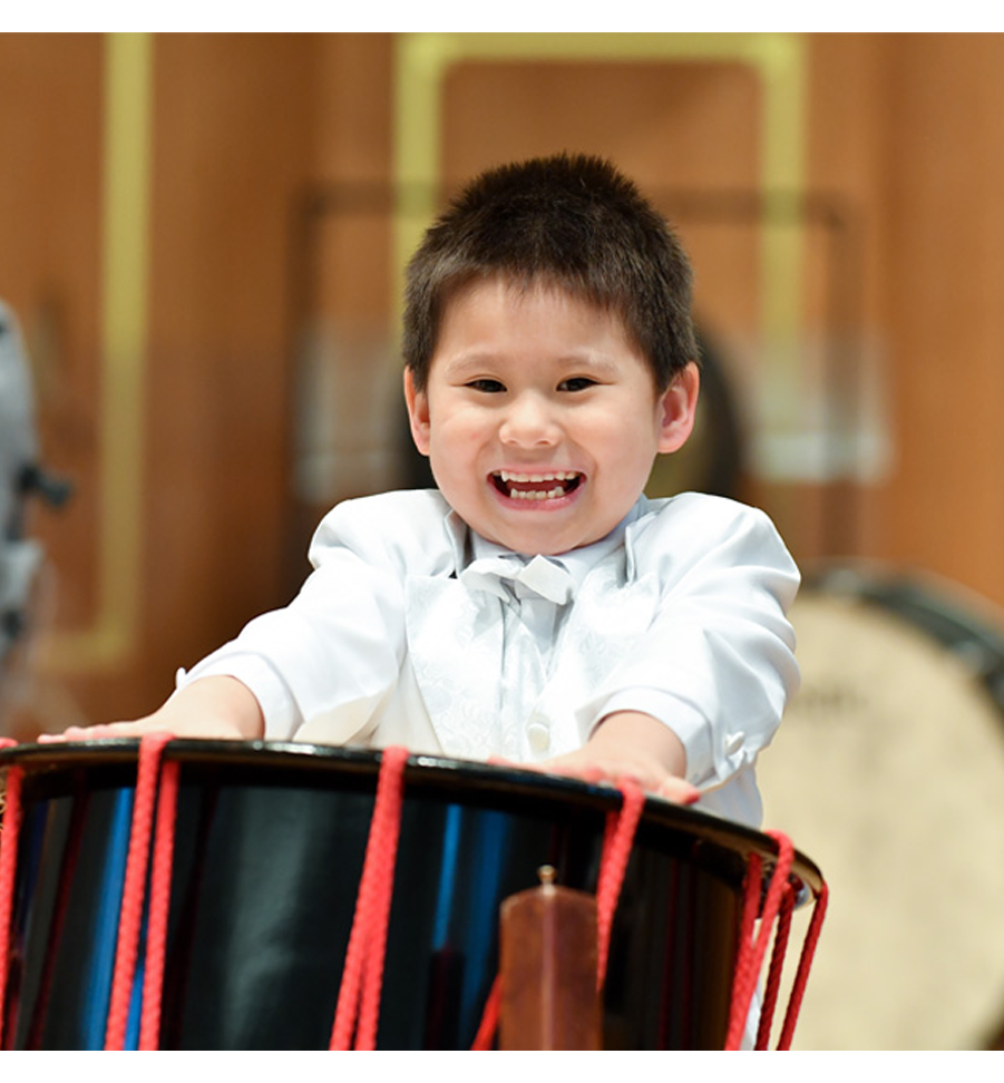 A young boy, wearing a white shirt, performing on a drum, smiling at the camera.